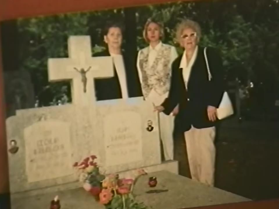 Three women standing beside two graves with a cross and flowers