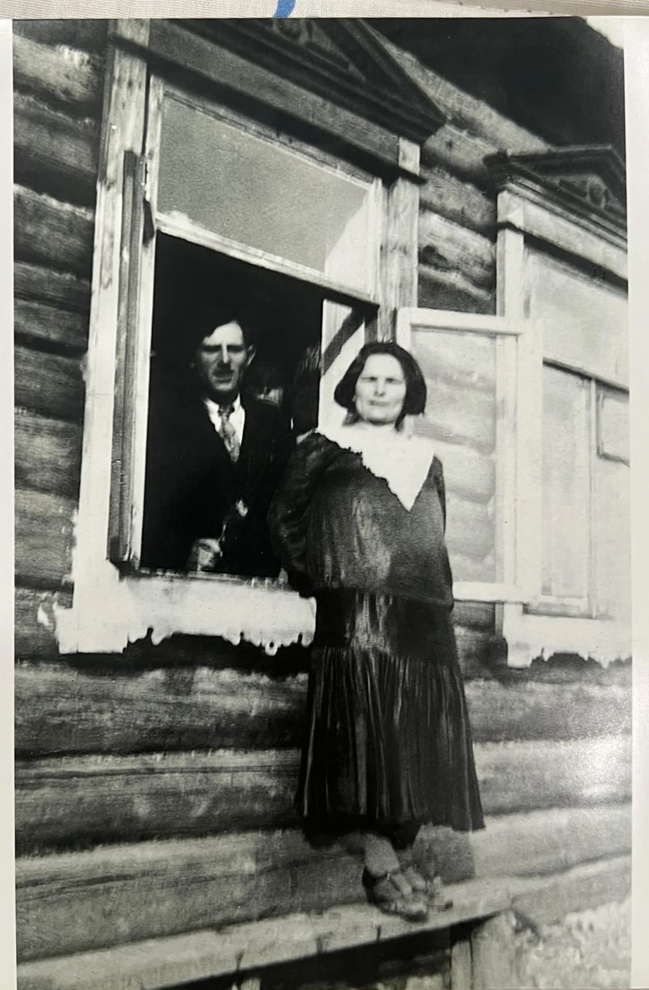 A man and a woman at the open window of a log house, the woman standing outside on the sill